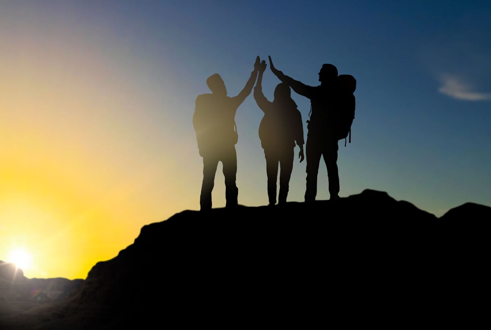 Siluetas de compañeros con mochilas chocando las manos en la cima de una montaña al atardecer, celebrando un logro de team building
