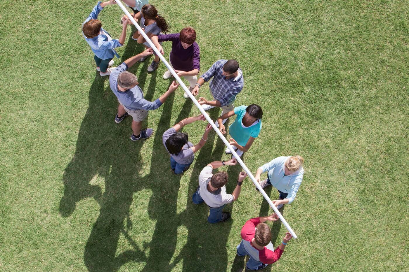 Vista aérea de un equipo de colaboradores sosteniendo juntos un tubo con la punta de los dedos sobre el pasto durante un reto de team building