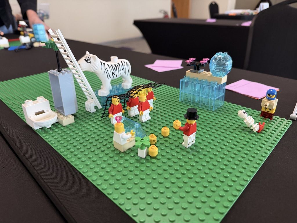 Escena construida con minifiguras, un tigre y piezas LEGO sobre una placa verde en la mesa de trabajo de un taller LEGO SERIOUS PLAY
